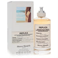 Maison Margiela Replica Beach Walk 3.4 oz EDT: elegant clear glass bottle with warm amber liquid, silver cap, and iconic white label, beside its minimalist box.