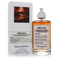 Replica By the Fireplace perfume bottle and box. Amber fragrance in a clear glass bottle with a white label and silver cap.